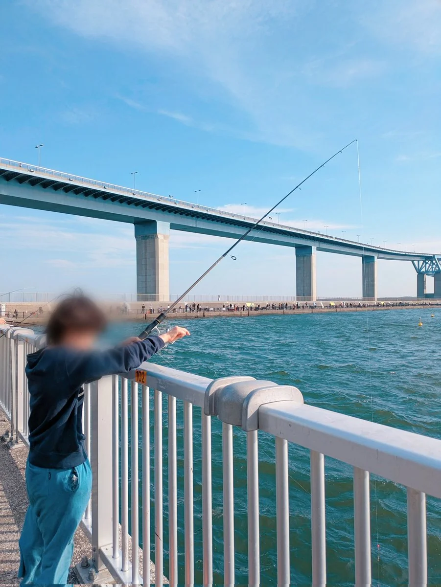 東京ゲートブリッジを背景に、海沿いの柵から釣り竿を伸ばす子ども